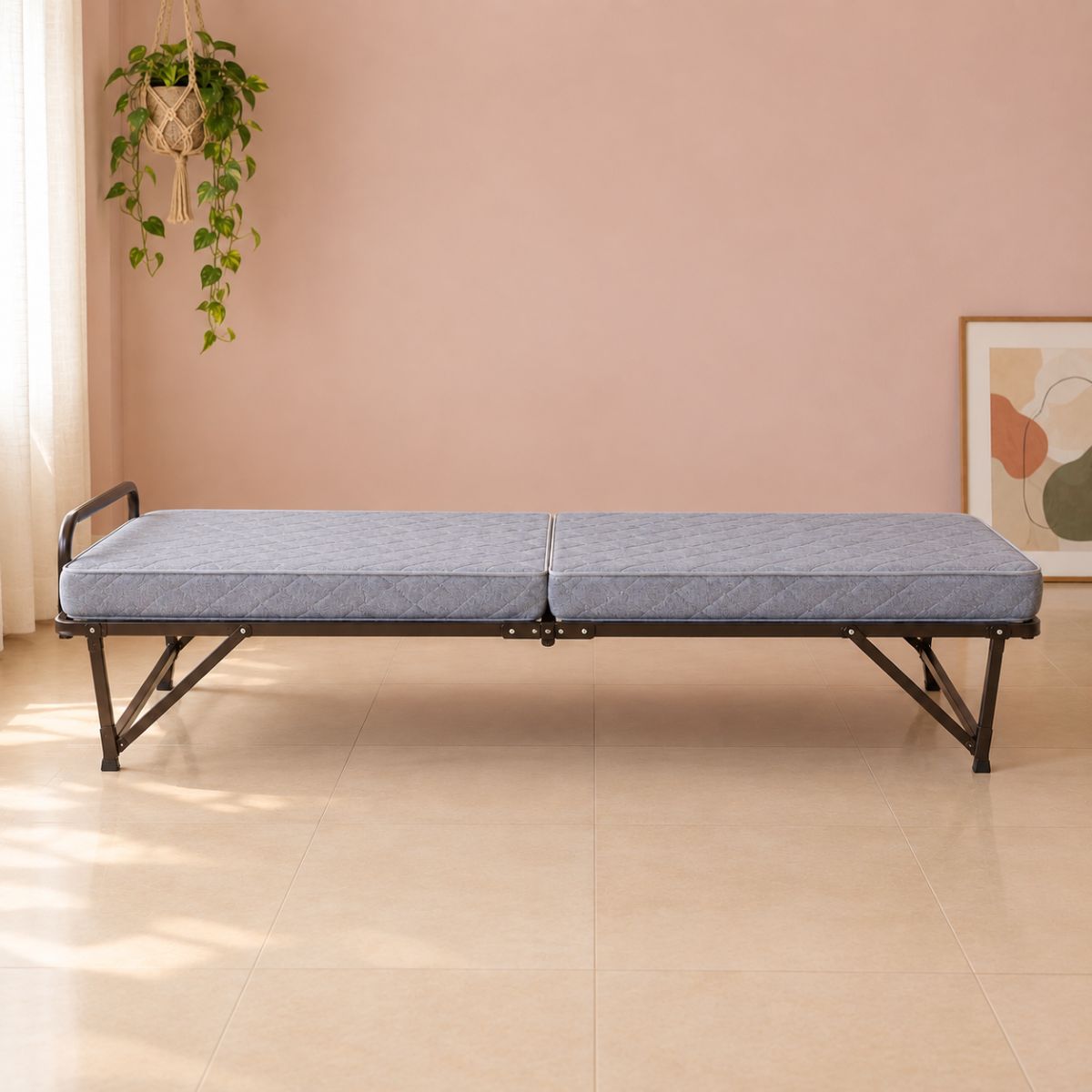 A fully extended folding bed with grey quilted mattress, dark metal angled legs and caster wheels against a blush pink wall